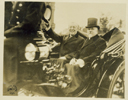 Black and white photograph of President Wilson, raising his hat in the air, and French President Poincare, sharing a carriage on a ride through the streets of Paris.