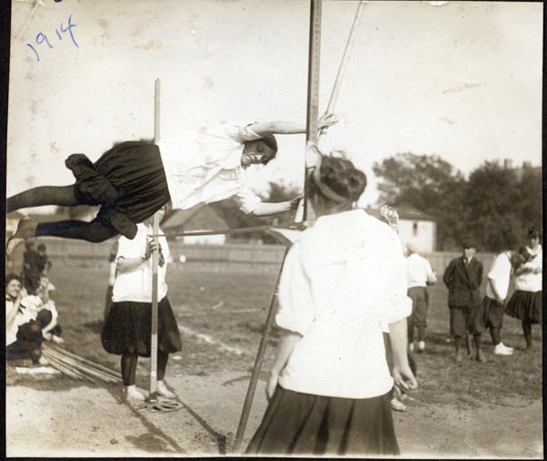 1914; black and white photograph of a woman pole vaulting, two women wait on either side