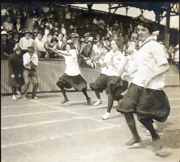 black and white photograph of five women at the finish-line of a race; man and woman stand at the line, near bleachers of onlookers
