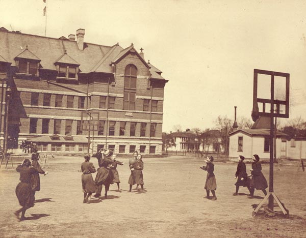 black and white photograph of women in all-dark uniforms playing outdoors; large building in background, basketball hoop in foreground